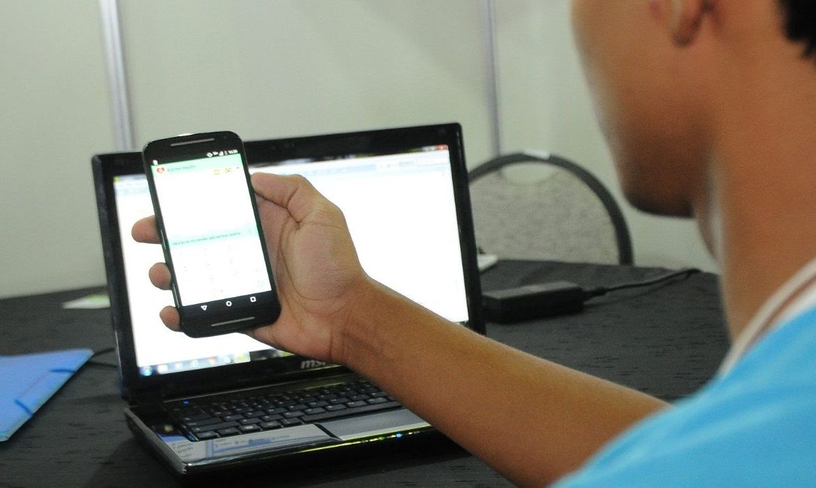 Aluno com celular na mão e um laptop no fundo - Internet Brasil