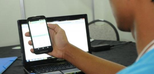 Aluno com celular na mão e um laptop no fundo - Internet Brasil